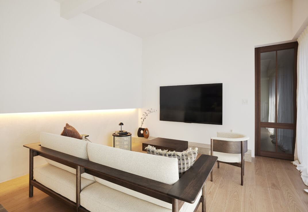 リビング。ソファとローテーブル、壁掛けテレビが並び、木製建具の奥に隣室がつながる。